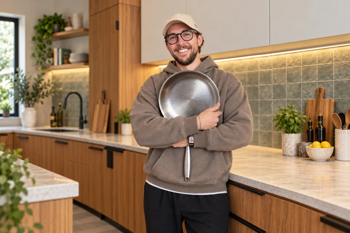 Smiling man in a cap hugging a Siraat titanium hammered frying pan in a bright modern kitchen